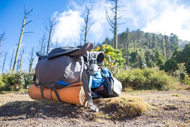 Large hiking backpack with gear and sleeping pad resting on the ground in a forested mountain area under a blue sky

