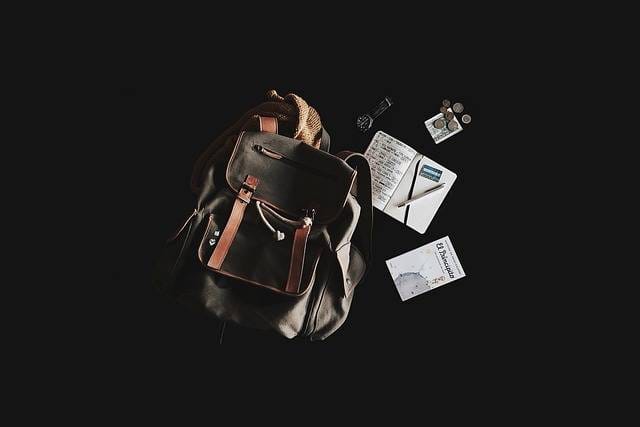Flat lay of a travel backpack with essentials including a notebook, passport, map, coins, and a pen on a dark background

