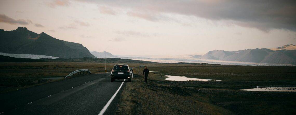 Scenic landscape at sunrise or sunset with a black car parked roadside and a person walking away toward snow-capped mountains under a partly cloudy sky, with small lakes dotting the terrain to the right.