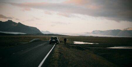 Scenic landscape at sunrise or sunset with a black car parked roadside and a person walking away toward snow-capped mountains under a partly cloudy sky, with small lakes dotting the terrain to the right.